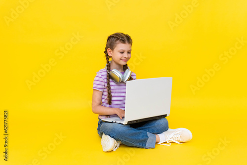 A young girl with a laptop. A little girl is sitting on the floor with her legs crossed and holding a laptop. Social networking for teenagers. Yellow isolated background.