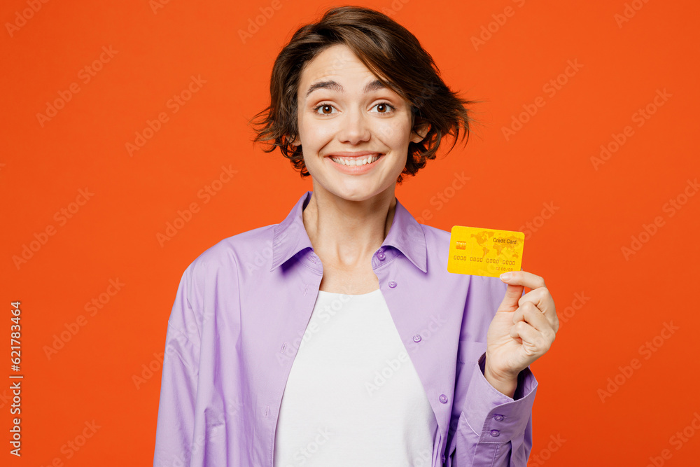 Young cheerful fun caucasian woman she wear purple shirt white t-shirt casual clothes hold in hand mock up of credit bank card isolated on plain orange background studio portrait. Lifestyle concept. Young cheerful fun caucasian woman she wear purple shirt white t-shirt casual clothes hold in hand mock up of credit bank card isolated on plain orange background studio portrait. Lifestyle concept.