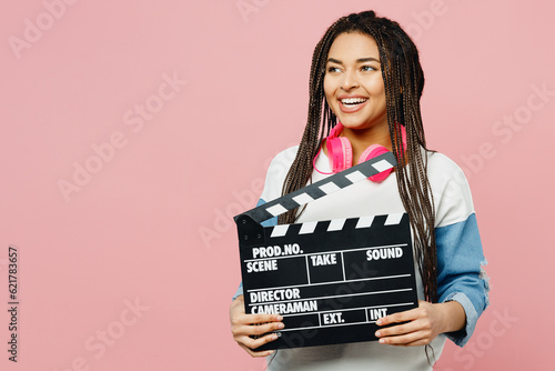 Young woman of African American ethnicity wears white sweatshirt casual clothes hold classic black film making clapperboard look aside isolated on plain pastel light pink background studio portrait.