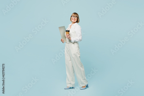 Full body employee IT business woman 50s wearing white classic suit glasses formal clothes holding closed laptop pc computer drink coffee isolated on plain blue background. Achievement career concept.