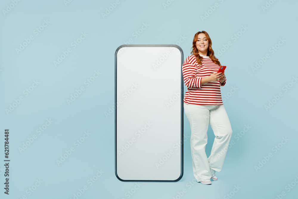 Full body smiling happy young chubby overweight woman wear red shirt casual clothes big huge blank screen mobile cell phone with area use smartphone isolated on plain blue background studio portrait. Full body smiling happy young chubby overweight woman wear red shirt casual clothes big huge blank screen mobile cell phone with area use smartphone isolated on plain blue background studio portrait.