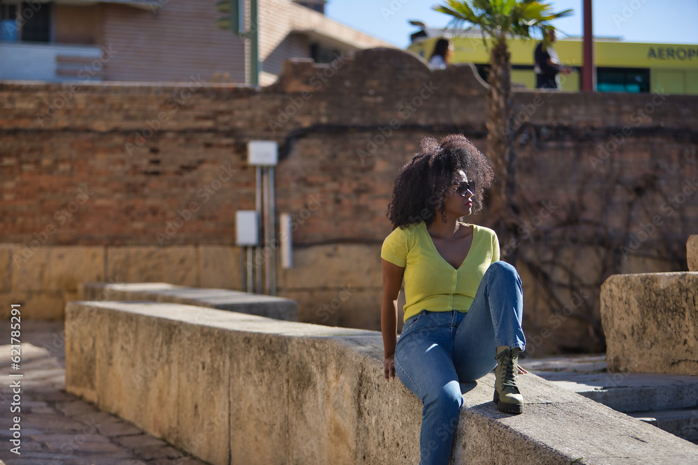 Young, beautiful, black woman with afro hair, wearing yellow t-shirt, jeans and sunglasses, sitting on a stone wall, relaxed, calm and sunbathing. Vacation concept, travel, current, modern.