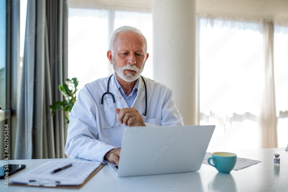 Serious mature doctor using laptop and sitting at desk. Senior professional medic physician wearing white coat and stethoscope working on computer at workplace. Serious mature doctor using laptop and sitting at desk. Senior professional medic physician wearing white coat and stethoscope working on computer at workplace.