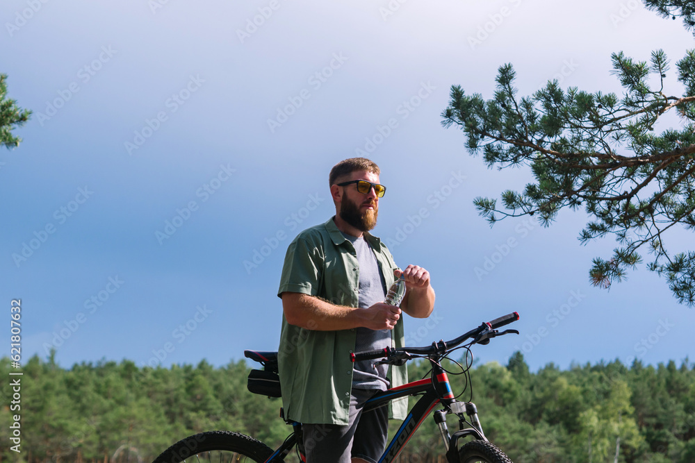 Fototapeta premium Cyclist have rest near the bicycle, in the forest and relax for energy in adventure workout, drinking bottle of water for hydration and enjoying in the forest trees for wellness. Bearded man on bike 