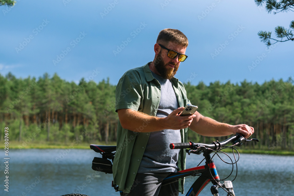 Obraz premium Happy bearded man in jeans clothes stand near bicycle bike on sidewalk in green park outdoors use mobile cell phone 13 Pro chat online. People active urban healthy lifestyle cycling concept