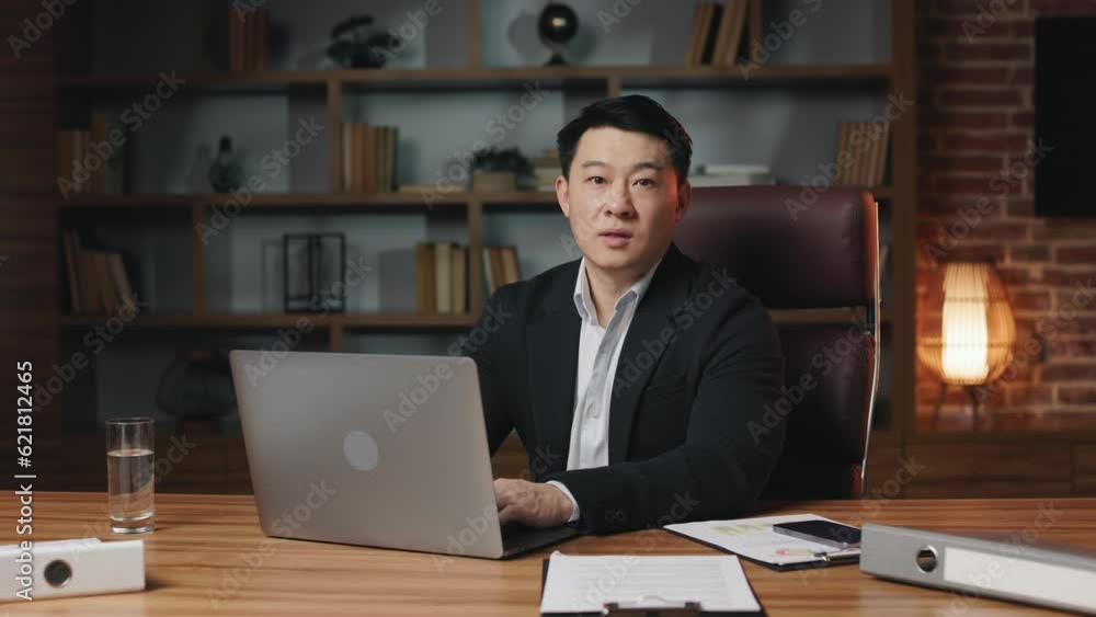 Ambitious male employee working in computer program using portable laptop and then looking at camera. Proactive asian manager wearing trendy business suit making gentle smile. Ambitious male employee working in computer program using portable laptop and then looking at camera. Proactive asian manager wearing trendy business suit making gentle smile.