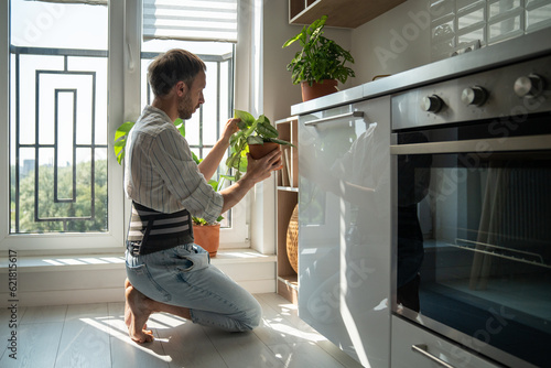 Young man wearing support belt taking care of houseplants at home, recovering after spinal injury, living with chronic back pain. Using orthopedic lumbar brace for pain relief