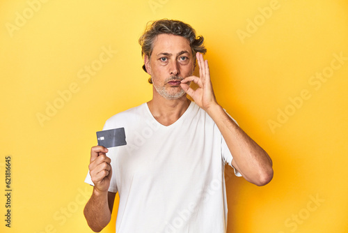 Man holding a credit card, yellow studio backdrop with fingers on lips keeping a secret. © Asier