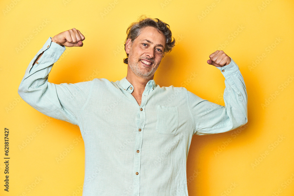 Fototapeta premium Middle-aged man posing on a yellow backdrop showing strength gesture with arms, symbol of feminine power