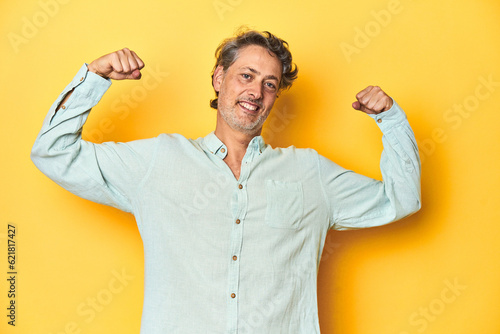 Middle-aged man posing on a yellow backdrop showing strength gesture with arms, symbol of feminine power