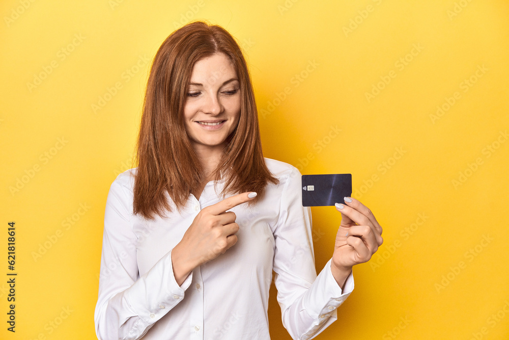 Redhead holding credit card, financial concept smiling and pointing aside, showing something at blank space.