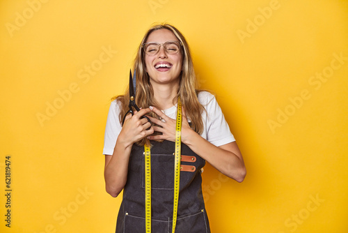 Stylish Caucasian stylist with scissors on yellow backdrop, laughs out loudly keeping hand on chest. © Asier