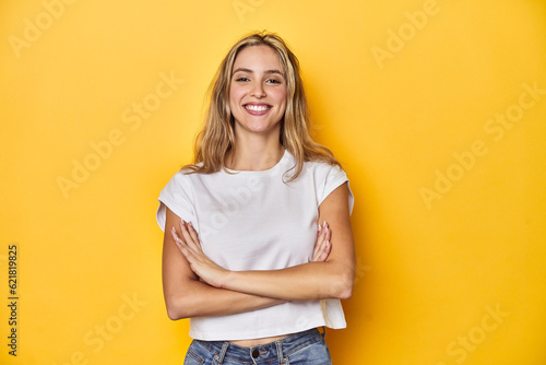 Young blonde Caucasian woman in a white t-shirt on a yellow studio background, who feels confident, crossing arms with determination.
