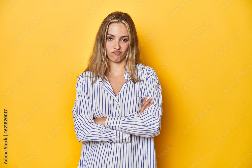 Young blonde Caucasian woman in a striped business shirt on a yellow background, frowning face in displeasure, keeps arms folded. Young blonde Caucasian woman in a striped business shirt on a yellow background, frowning face in displeasure, keeps arms folded.
