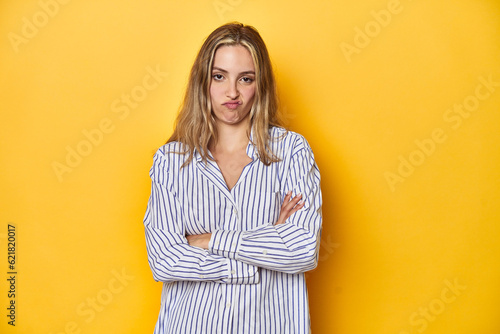 Young blonde Caucasian woman in a striped business shirt on a yellow background, frowning face in displeasure, keeps arms folded.