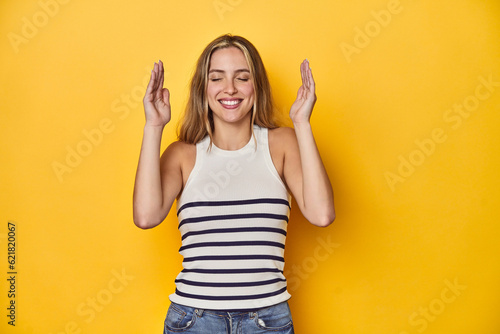 Young blonde Caucasian woman in a white tank top on a yellow studio background, joyful laughing a lot. Happiness concept.