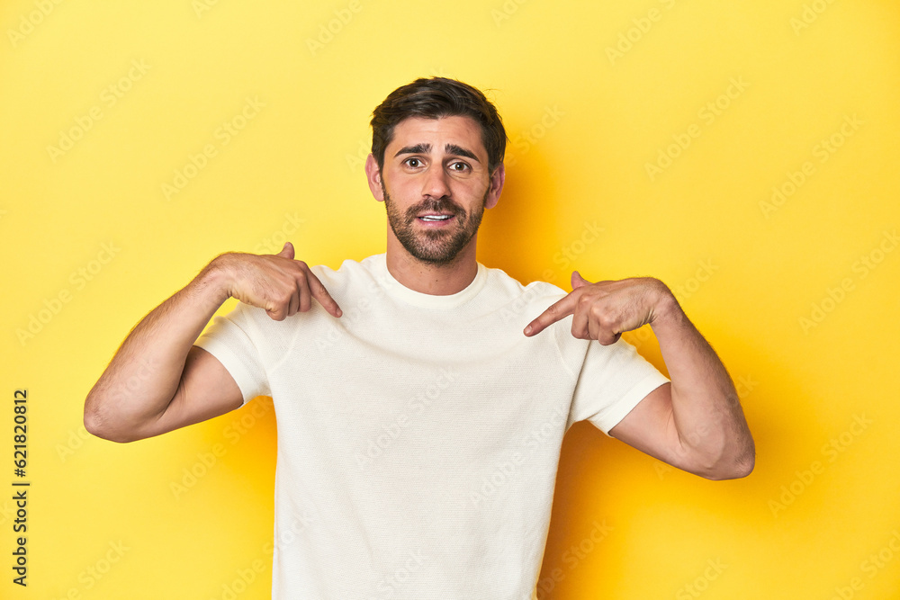Caucasian man in white t-shirt on yellow studio background surprised pointing with finger, smiling broadly. Caucasian man in white t-shirt on yellow studio background surprised pointing with finger, smiling broadly.