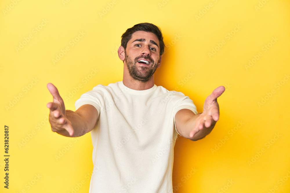 Caucasian man in white t-shirt on yellow studio background feels confident giving a hug to the camera. Caucasian man in white t-shirt on yellow studio background feels confident giving a hug to the camera.