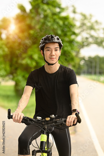 Smiling young asian man dressed in casual clothes and helmet riding a bicycle along the bike path in a city park