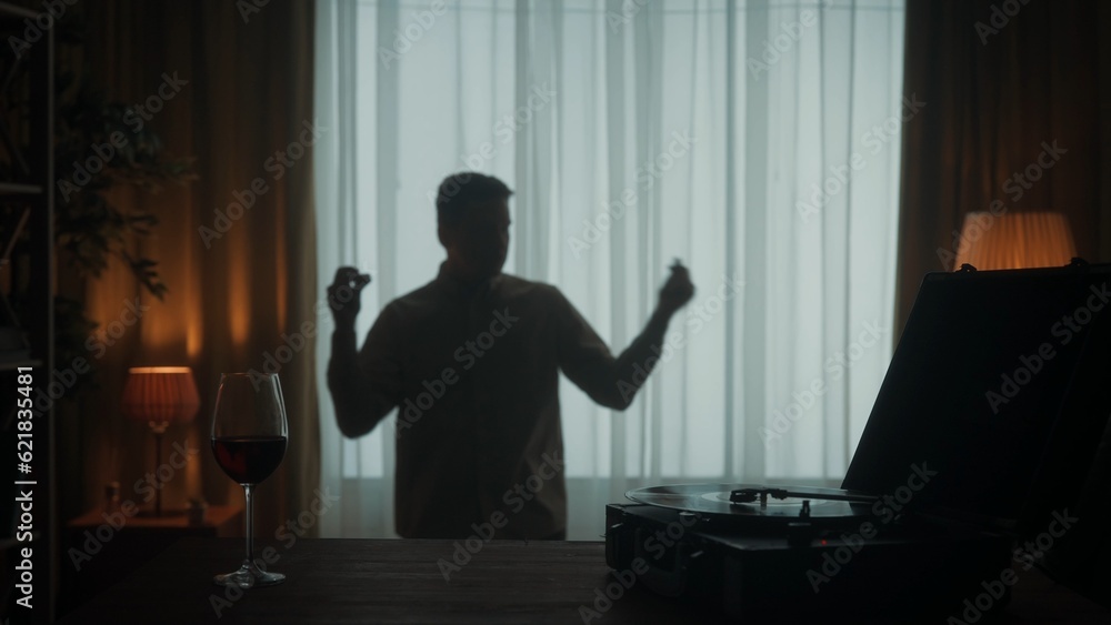 Vintage record player with a rotating vinyl record and a glass of red wine on the table close up. A carefree person dances merrily alone. The dark silhouette of a dancing man in the living room.