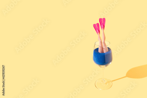 Female legs with pink flippers in blue sand sticking out of a drinking glass on a bright yellow background. Summer beach enjoyment concept.