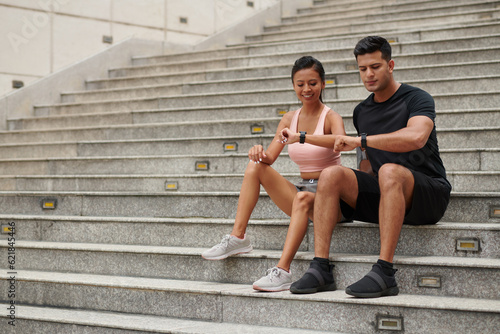 Smiling couple sitting on steps and checking fitness trackers after morning jog