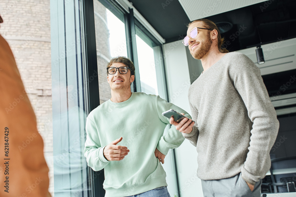Fototapeta premium smiling entrepreneur in stylish eyeglasses holding mobile phone while standing with hand in pocket near business team and large windows in coworking office, concept of successful collaboration