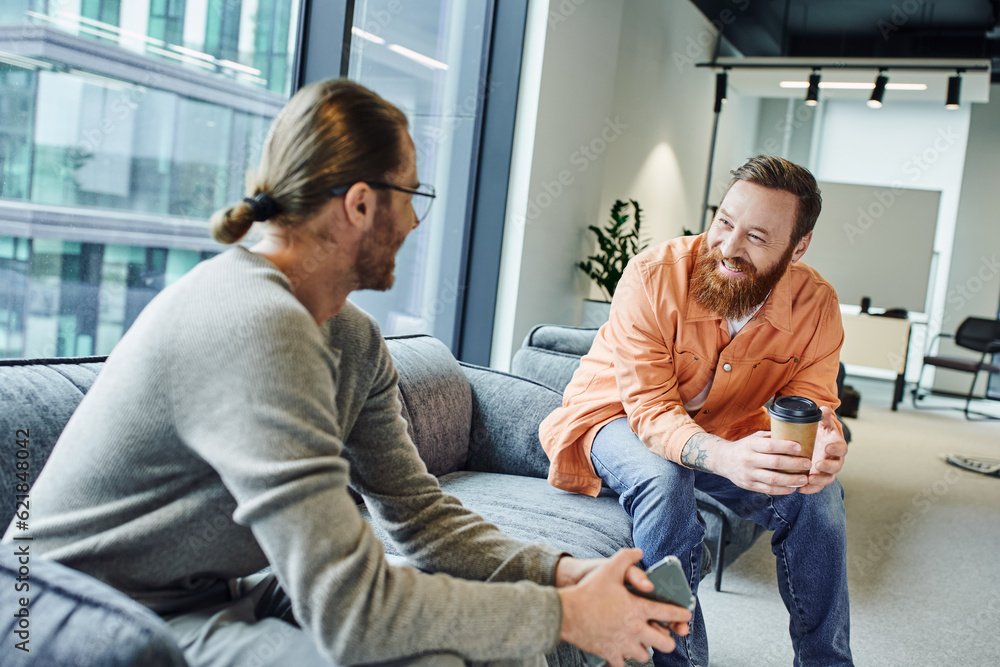 Obraz premium cheerful, bearded and tattooed businessman with coffee to go discussing startup project with colleague sitting with mobile phone on couch in office lounge of modern coworking environment
