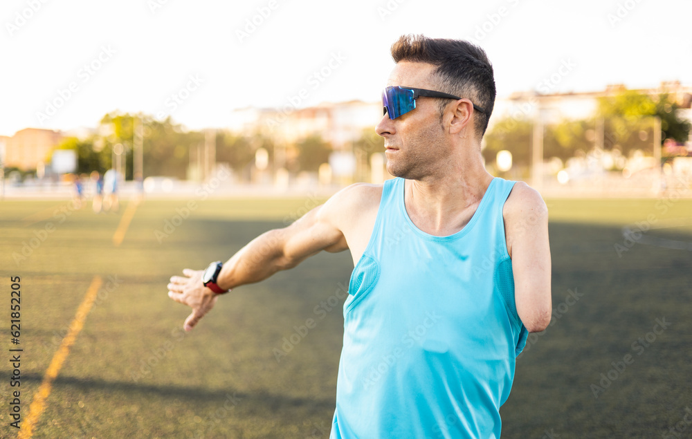 An adult senior soccer player stretches out an arm in the middle of a soccer field. The man is wearing sports glasses and a tank top. Concept of disabled footballers, athletes with an amputated arm. An adult senior soccer player stretches out an arm in the middle of a soccer field. The man is wearing sports glasses and a tank top. Concept of disabled footballers, athletes with an amputated arm.