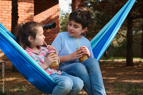 Happy children, teen boy and his younger sister, drinking fresh fruit smoothie while relaxing on hammock in the backyard