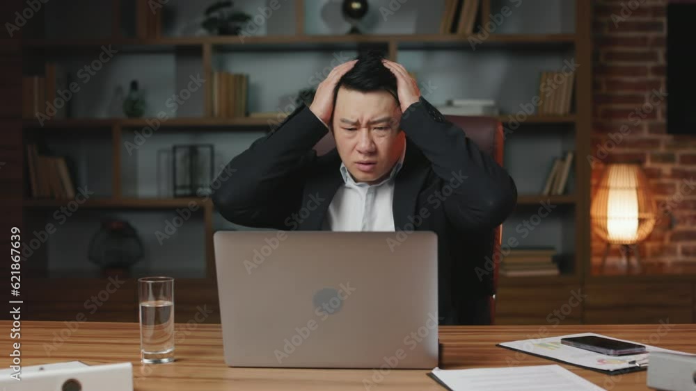 Front view of overwhelmed asian chief executive reading horrible news in internet while sitting at office desktop with portable computer. Perplexed man feeling shock and catching head with hands. Front view of overwhelmed asian chief executive reading horrible news in internet while sitting at office desktop with portable computer. Perplexed man feeling shock and catching head with hands.