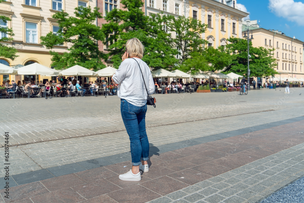 Chic blonde woman, wearing blue jeans and a white shirt, confidently stands on the sidewalk, phone in hand. In the background, a sun-soaked summer restaurant buzzes with people seated at tables
