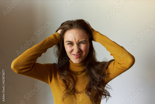 Horizontal portrait of annoyed frowning scowling grimacing female of 20s pulling her hair being in bad mood, looking at camera against gray studio background in yellow turtleneck sweater photo
