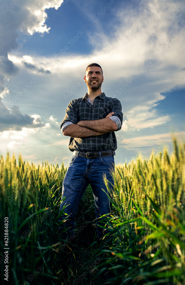 Portrait of young farmer standing in a green wheat field at sunset.