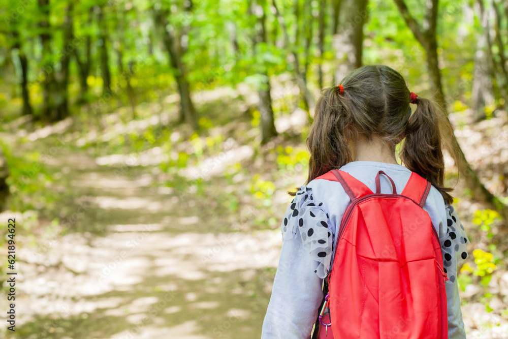 Little girl with backpack walking on trail in the spring forest. Back view.
