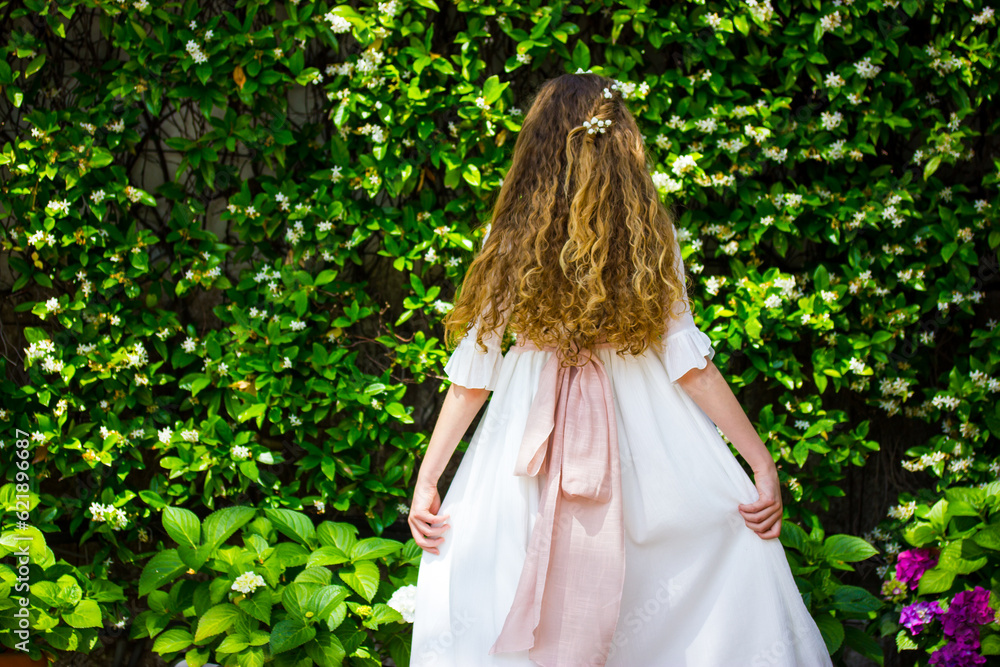 Little girl in a long white dress with beautiful long curly hair view from a back against a background of flowering shrubs. A princess in a spring garden. Wedding female fashion. Faceless woman, lady. Little girl in a long white dress with beautiful long curly hair view from a back against a background of flowering shrubs. A princess in a spring garden. Wedding female fashion. Faceless woman, lady.
