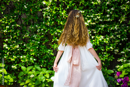 Little girl in a long white dress with beautiful long curly hair view from a back against a background of flowering shrubs. A princess in a spring garden. Wedding female fashion. Faceless woman, lady.