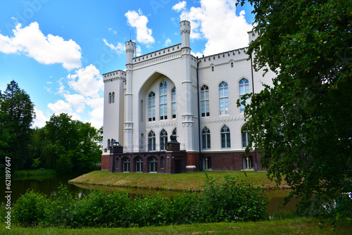 Old castle with white walls, arch and high windows. Green trees all around and blue sky. Natural historical park. Poland, Kurnik castle, Poznan, June 2022.