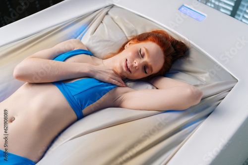 Top side view of relaxed woman wearing bikini lying with closed eyes on electric massage bed, having rest enjoying spa procedure at luxury spa salon. Sensual redhead female enjoying wellness weekend. photo