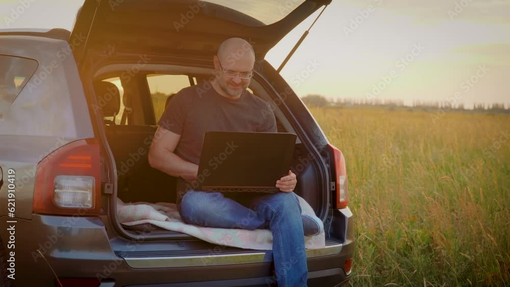 Smiling male freelancer working on laptop, man typing on keyboard, sitting in car trunk while traveling, close up. Busy worker freelancer entrepreneur notebook online working outdoors in SUV Smiling male freelancer working on laptop, man typing on keyboard, sitting in car trunk while traveling, close up. Busy worker freelancer entrepreneur notebook online working outdoors in SUV