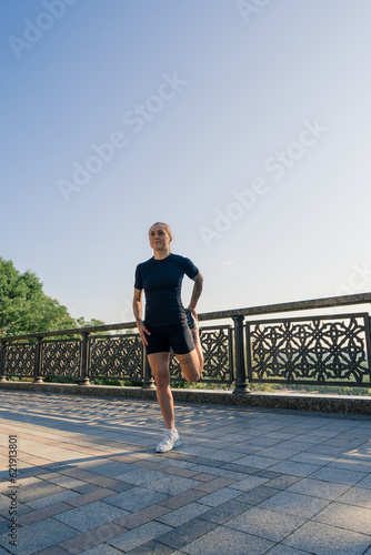 Fitness young sportswoman girl in the morning doing warm-up leg exercises before training on the street in the city on the bridge Sport health