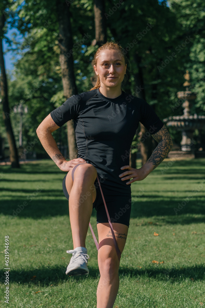 Obraz premium young sportswoman girl doing physical exercises on a sports mat training with rubber bands on her legs outside in the park healthy lifestyle concept