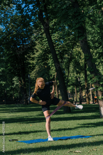 young sportswoman girl doing physical exercises on a sports mat training with rubber bands on her legs outside in the park healthy lifestyle concept