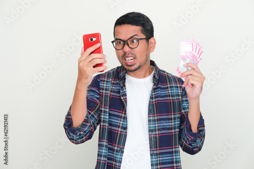 A man looking to his mobile phone with angry expression with one hand holding money