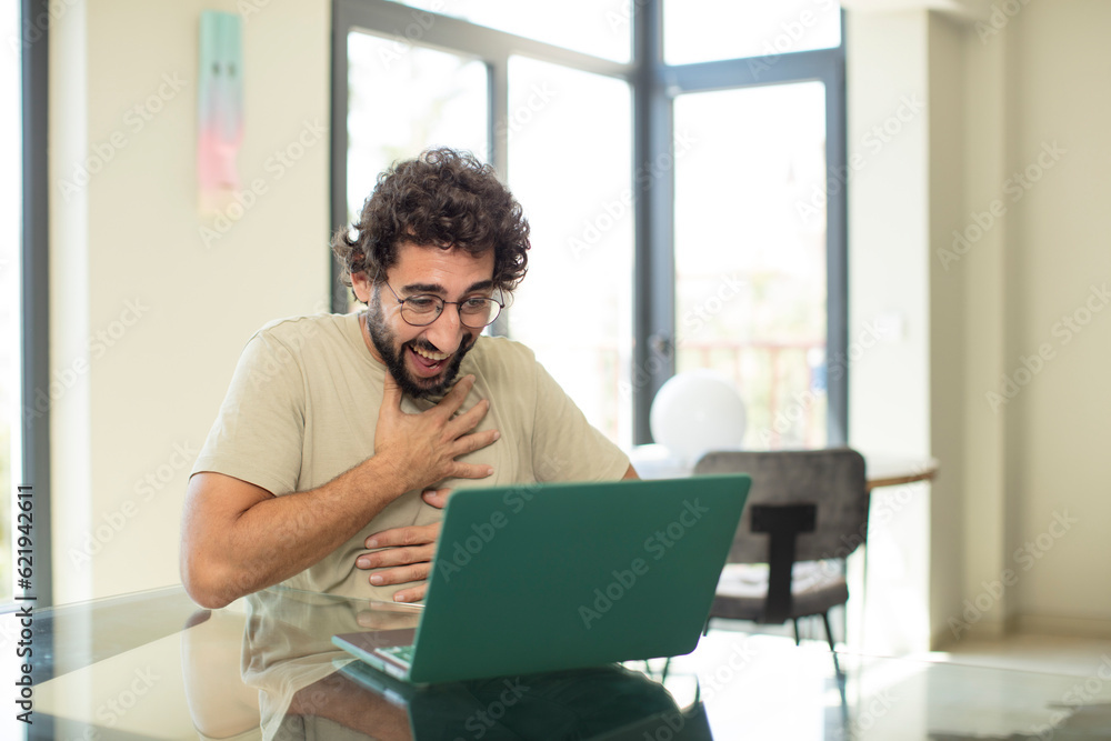 young adult bearded man with a laptop laughing out loud at some hilarious joke, feeling happy and cheerful, having fun