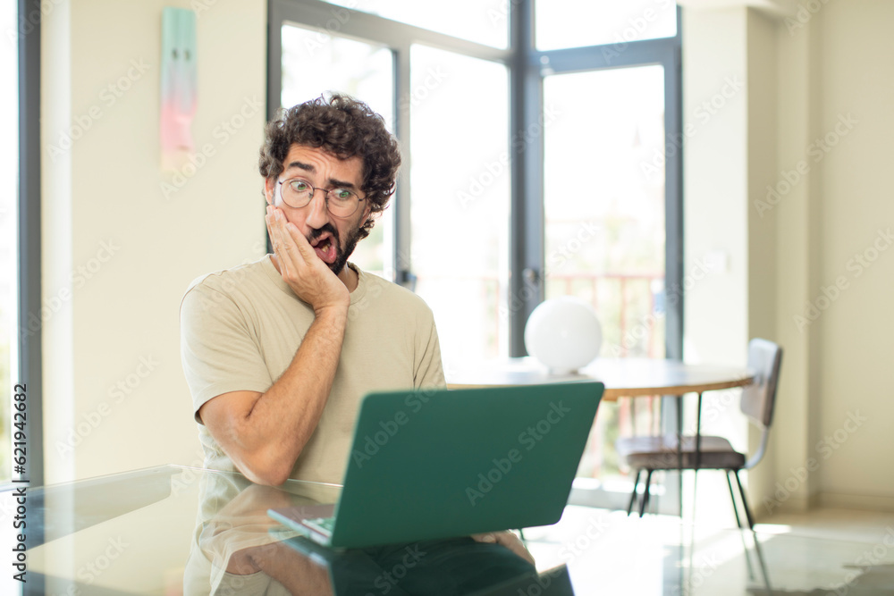 young adult bearded man with a laptop feeling shocked and astonished holding face to hand in disbelief with mouth wide open young adult bearded man with a laptop feeling shocked and astonished holding face to hand in disbelief with mouth wide open