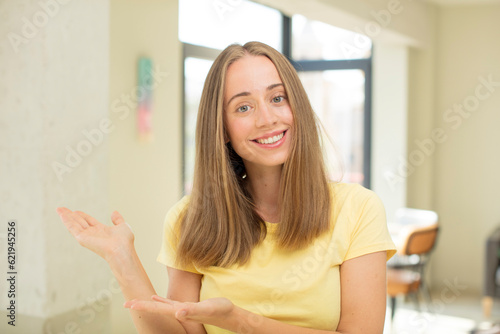 pretty blond woman feeling happy and cheerful, smiling and welcoming you, inviting you in with a friendly gesture