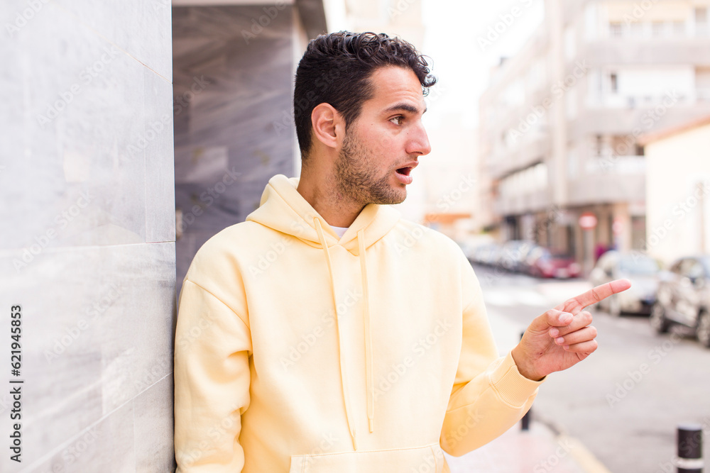 Obraz premium young hispanic man feeling shocked and surprised, pointing and looking upwards in awe with amazed, open-mouthed look