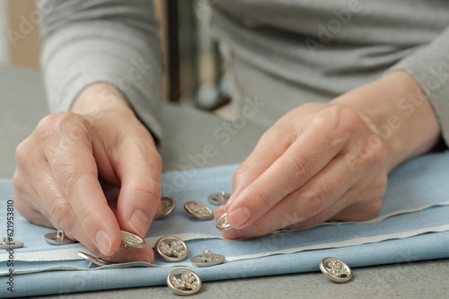 Production of clothing in a sewing studio. A tailor woman picks up sewing buttons for a future sewing product. Metal buttons and blue textile material are scattered on the desktop.