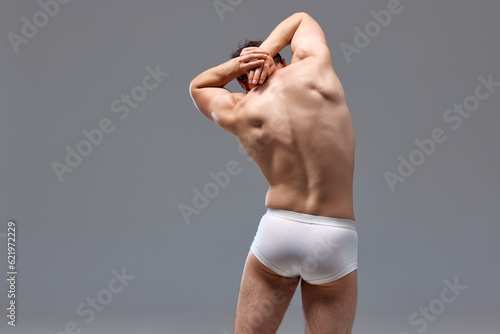 Rear view of muscular, relief male back. Man posing shirtless in underwear against grey studio background. Stretching. Concept of man's beauty, sport, health, athletic body, medicine photo